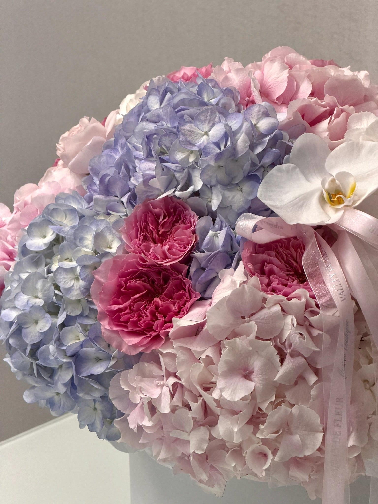 Birthday floral arrangement featuring light blue and pink hydrangeas, deep pink carnations, a white orchid, and a pale pink ribbon bow.